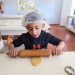 Un petit garçon avec une charlotte sur la tête et un rouleau à pâtisserie étale un sablé