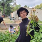 Un jeune homme présente des pommes de terres fraichement récoltées. En arrière plan deux jeunes récoltent les pommes de terre dans le potager