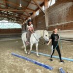 Une jeune fille sur un cheval blanc effectue un parcours. devant elle, une autre jeune fille tient la bride du cheval