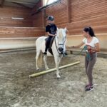 Une jeune fille sur un cheval blanc qui enjambe un obstacle au sol. Le cheval est tenu par la bride par une animatrice