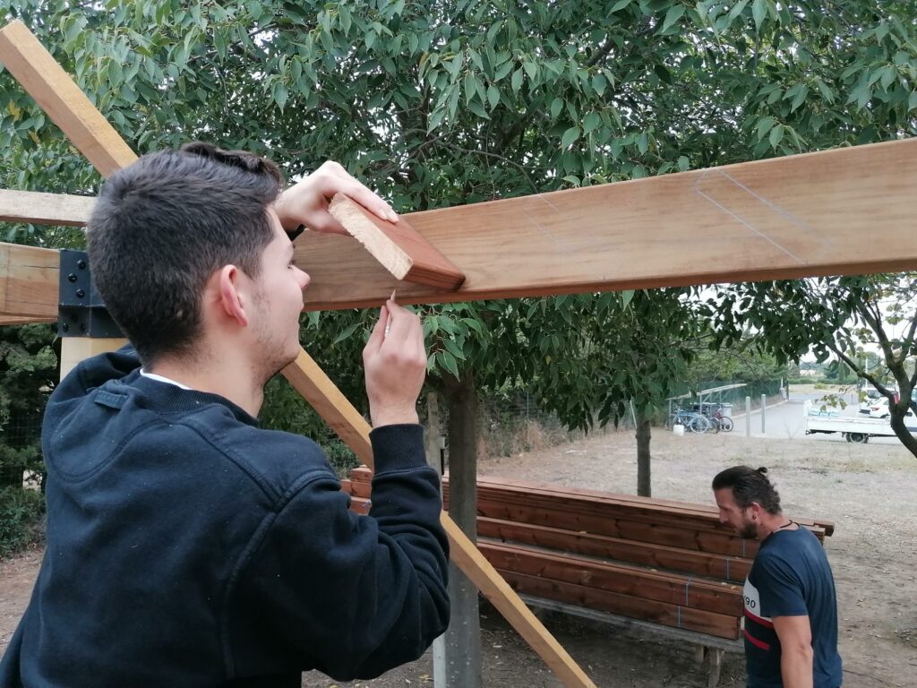 Un jeune, de dos, travaille sur une pergola en bois. Au second plan, un adulte.