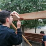 Un jeune, de dos, travaille sur une pergola en bois. Au second plan, un adulte.