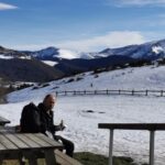 Une personne assise à une table de camping en bois devant un panorama de montagnes enneigées